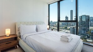 A modern hotel room with a large bed, a nightstand, a lamp, and a view of the cityscape through large windows. - High Floor River View 1BR Prime Location w/Parking (South Brisbane)