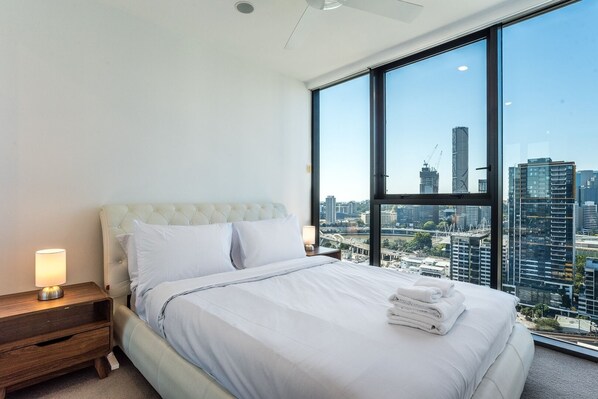 Quarto de hotel moderno com uma cama grande, uma mesa de cabeceira, uma luminária e vista para a cidade através de amplas janelas.