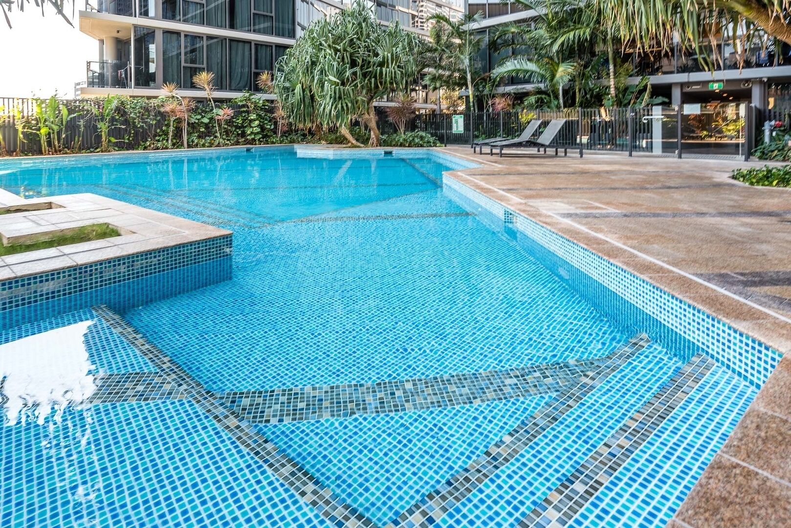 A swimming pool with tiled edges, surrounded by a paved area with lounge chairs and a building in the background.