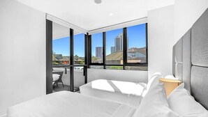 A modern hotel room with a large bed, a view of the city through floor-to-ceiling windows, and a balcony with a chair. - Cozy and Bright 2-bedroom Apartment in Surfers (Surfers Paradise)