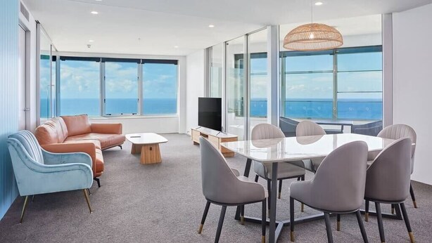 A modern living room with a glass table, grey chairs, a brown sofa, and a television.