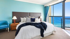 Una habitación de hotel con una cama grande, dos mesitas de noche con lámparas, un sillón azul y vista al océano.