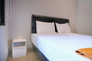 A neatly made bed with white bedding and a black headboard, a small white nightstand with a lamp, and a beige towel on the bed. - Djuragan Kamar Balawan (Malang)