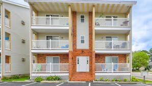 A multi-story apartment building with a central brick section, white balconies, and a parking lot in front. - Spacious 2 Bedroom Condo in North Ocean City! (Ocean City)