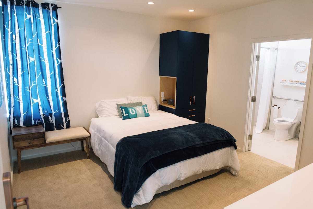 A bedroom with a bed, a bench, a wardrobe, and a bathroom visible through an open door.