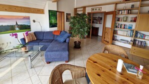 Un salon comprenant un canapé d’angle, une plante en pot, une bibliothèque et un coin repas avec une table en bois.