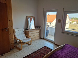 A bedroom with a wooden dresser, a white chair, a purple bed, a large window, and a potted plant. - Apartment 'Storchenblick' with Mountain View, Balcony, and Wi-Fi (Raisting)