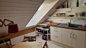 Une cuisine avec un plafond en pente, des meubles en bois, une étagère intégrée où sont rangés des bocaux, et une fenêtre.
