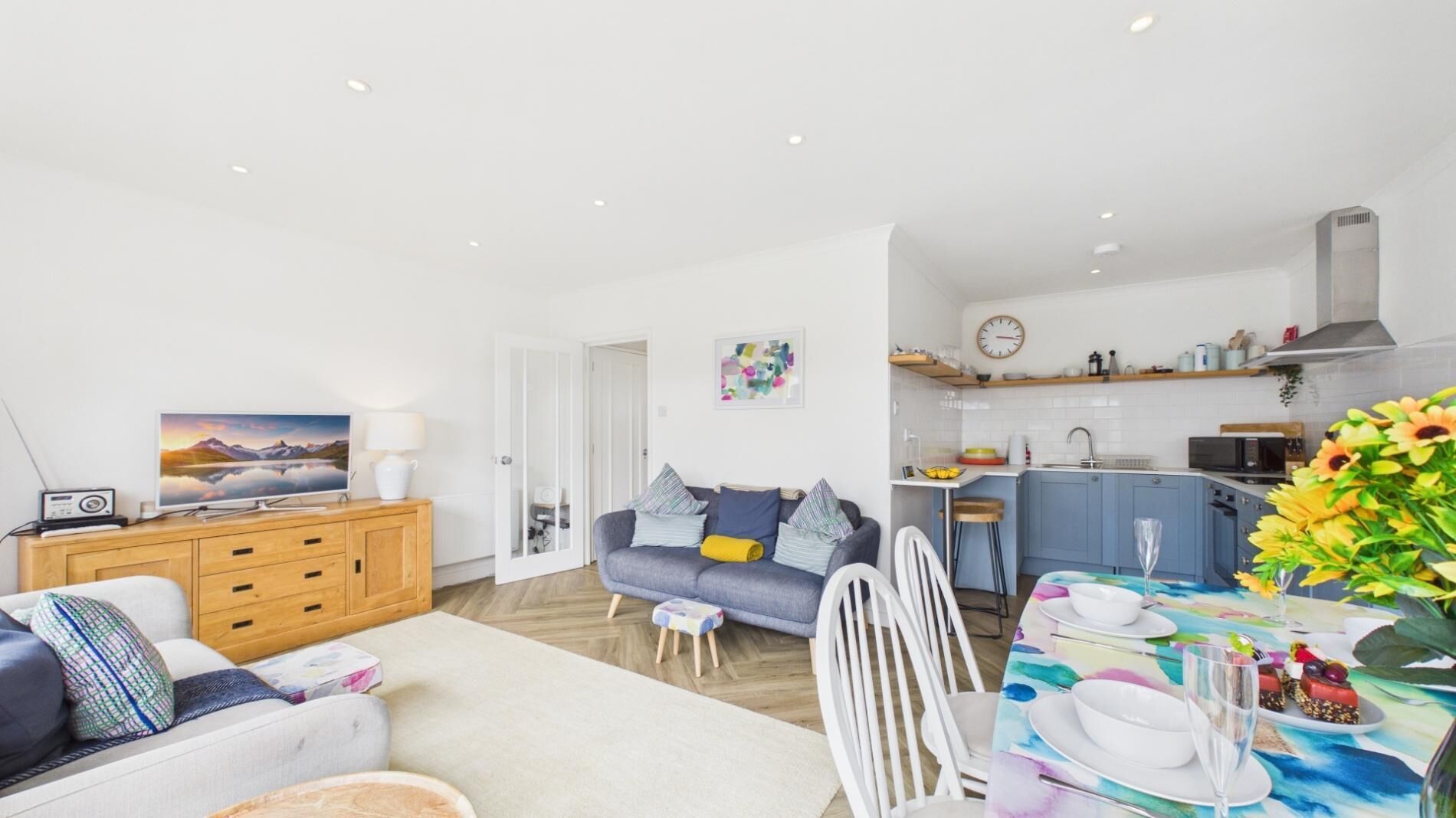 A modern living room with a sofa, a dining table set for two, and a kitchen area with a microwave and sink.