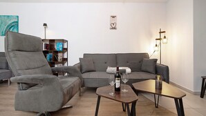 A modern living room with a grey sofa, a recliner, a coffee table with wine glasses, and a bookshelf.
