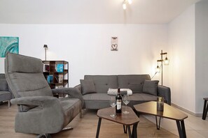 A modern living room with a grey sofa, a recliner, a coffee table with wine glasses, and a bookshelf.