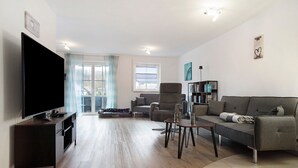 A modern living room with a flat-screen TV, grey sofa, armchairs, a coffee table, and a bookshelf.