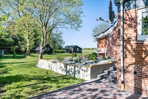 A brick building with a satellite dish, a concrete planter with plants, and a grassy yard with trees. - Tiny Home 'Vogelkoje' with Shared Terrace, Shared Garden and Wi-Fi (Emmelsbüll-Horsbüll)