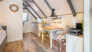 A cozy room with a wooden ceiling, a dining table with chairs, a small kitchen area, and a bed with a white headboard.