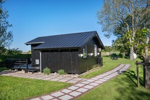A small, dark wooden building with a sloped roof, a paved pathway leading to it, and a grassy area with trees and a bench. - Holiday Home 'Achtern Diek' with Shared Terrace, Shared Garden and Wi-Fi (Emmelsbüll-Horsbüll)