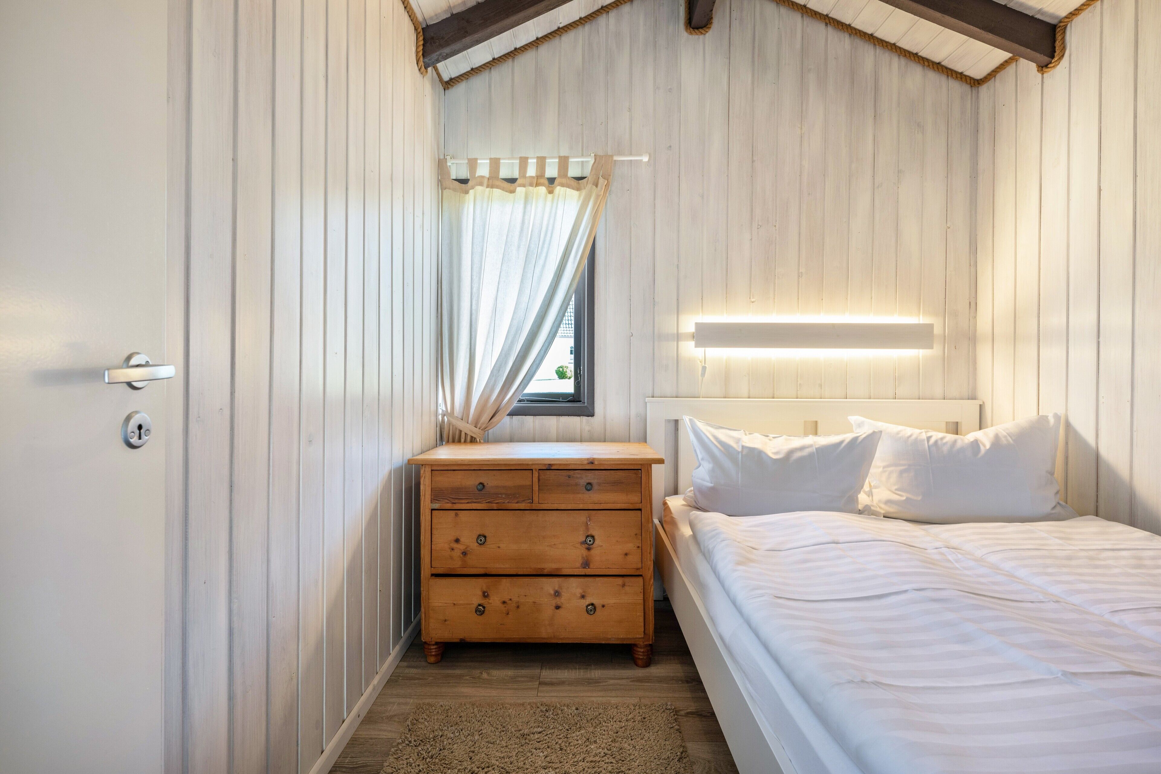 A bedroom with a wooden dresser, a bed with white bedding, a nightstand, and a window with curtains.