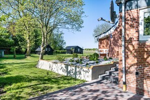 A brick building with a satellite dish, a concrete planter with plants, and a grassy yard with trees. - Holiday Home 'Achtern Diek' with Shared Terrace, Shared Garden and Wi-Fi (Emmelsbüll-Horsbüll)