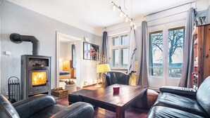 A cozy living room with a wood-burning stove, leather furniture, a dining table, and a view through large windows. - Holiday Home 'Seeblick' with Lake View, Private Terrace and Wi-Fi (Rheinsberg)