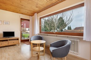Une pièce avec un plafond en bois, une grande fenêtre donnant sur des arbres, un téléviseur posé sur un meuble en bois et deux chaises.