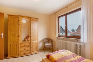 Une chambre avec une armoire en bois, une chaise en osier, un lit recouvert d’une couvre-lit à motifs et une fenêtre donnant sur des immeubles.