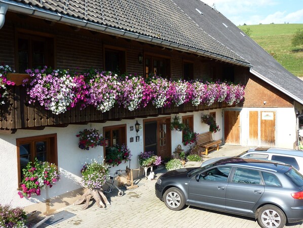 A traditional building with a balcony adorned with hanging flower baskets, a wooden bench, and a parked car. - Room 'Gästezimmer' with Private Terrace, Shared Garden and Wi-Fi (Breitnau)