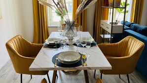 Ein Esstisch mit Tellern, Gläsern und Besteck, an beiden Seiten zwei Stühle, in der Mitte eine Vase mit Trockenpflanzen.