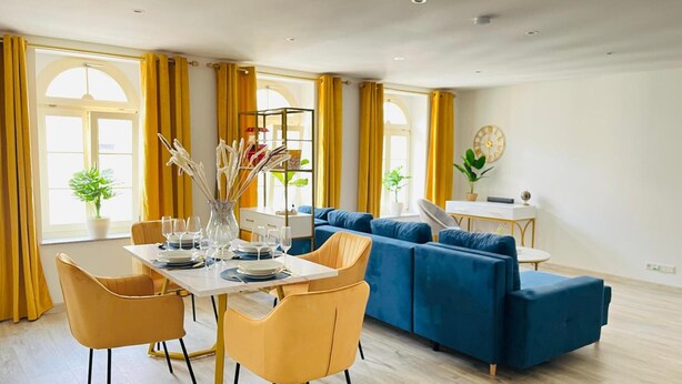 Ein modernes Wohnzimmer mit Essbereich, einem blauen Sofa, gelben Sesseln und einem Esstisch für sechs Personen.