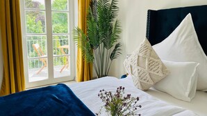 Ein Schlafzimmer mit Bett, einem Fenster mit Vorhängen, einer Pflanze und Blick ins Freie.