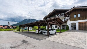 Ein überdachter Parkbereich mit zwei Autos, ein Gebäude mit Holzfassade und ein Berg im Hintergrund.