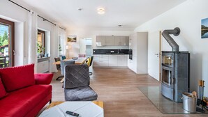 Ein modernes Interieur mit einem roten Sofa, einem grauen Sessel, einem Glastisch und einer Küche mit weißen Schränken sowie einem Kaminofen.