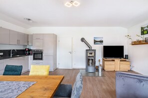 Une cuisine moderne avec des meubles blancs, un coin repas comprenant une table et des chaises en bois, un canapé gris, un téléviseur et un petit îlot de cuisine doté d’un évier.