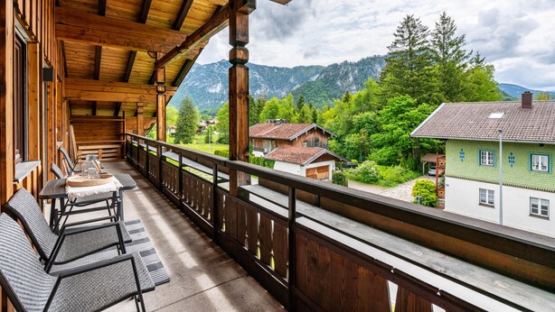 Ein hölzerner Balkon mit Sitzgelegenheit im Freien, Blick auf einen Berg und traditionelle Häuser im Hintergrund.