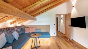 Une pièce moderne avec un plafond en bois, un canapé bleu agrémenté de coussins, une petite table ronde et un téléviseur fixé au mur.