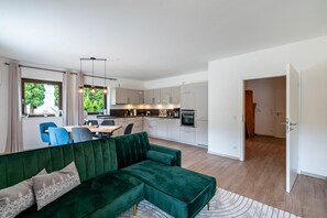 Un salon moderne avec un canapé d’angle vert, un coin repas avec des chaises bleues, une cuisine avec des meubles blancs et une vue par la fenêtre.