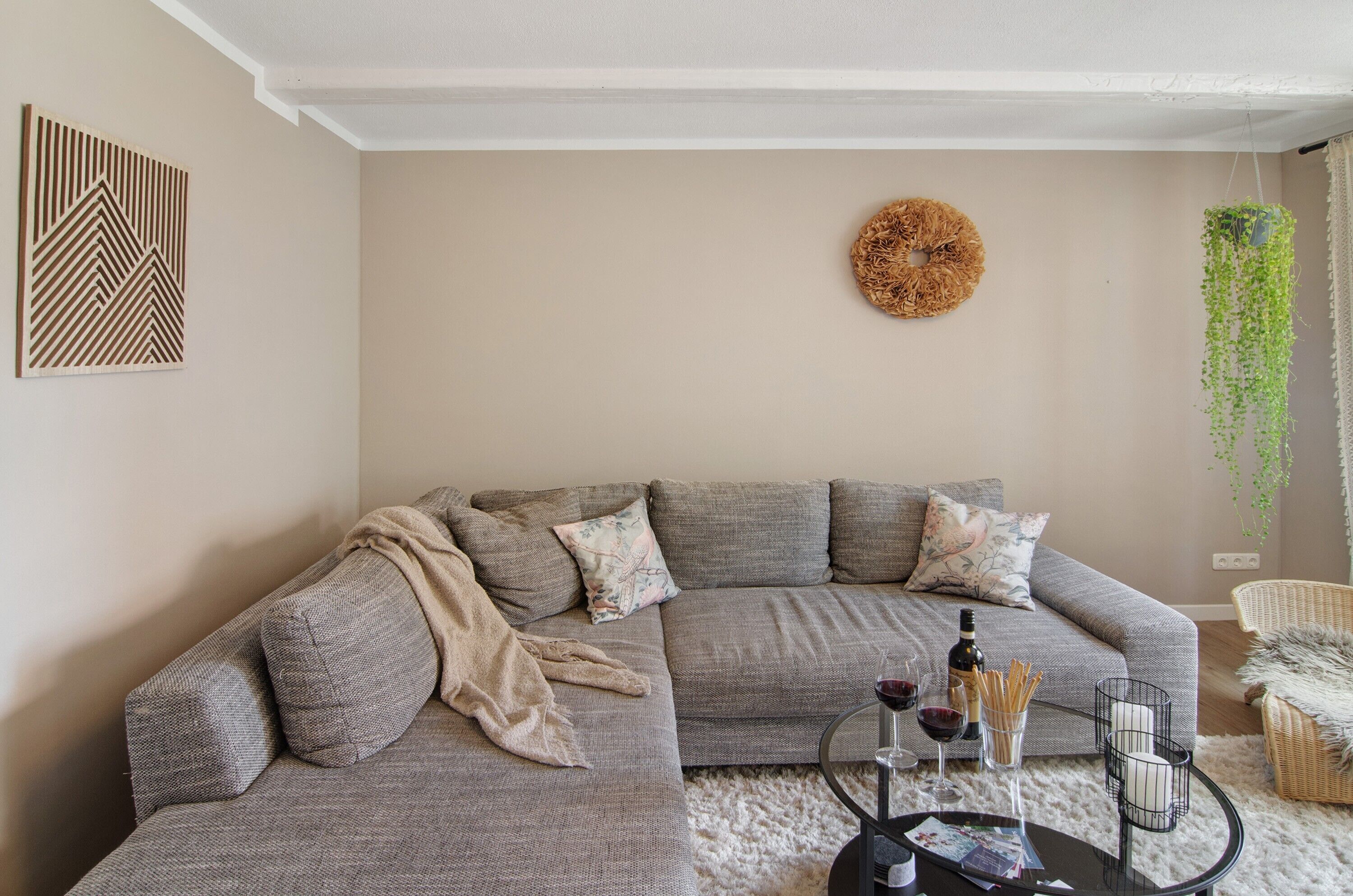 Un salon avec un canapé d’angle gris, un élément décoratif rond fixé au mur, une plante suspendue et une petite table sur laquelle se trouvent une bouteille et des verres.