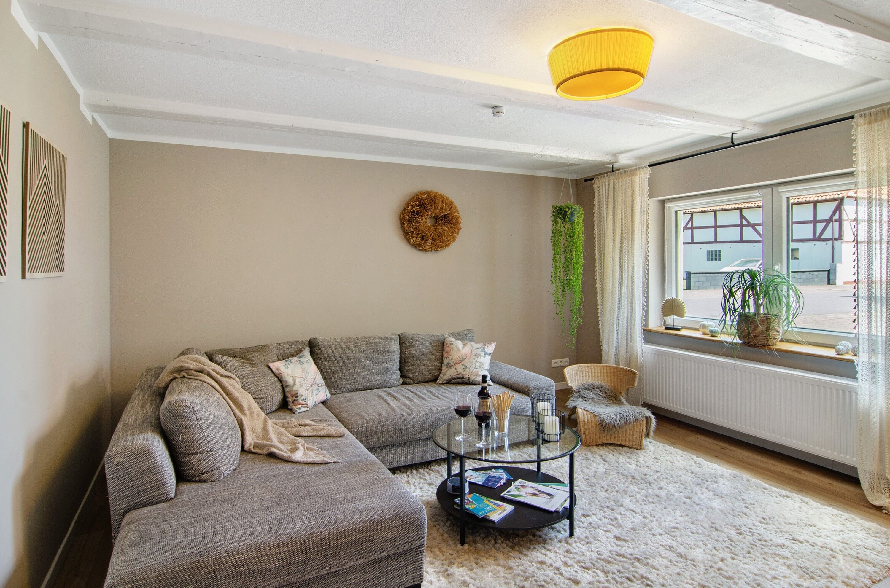 Una sala de estar con un sofá seccional gris, una mesa redonda de centro con libros y una vela, una planta colgante y un ventanal con vista a un edificio.