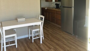 A compact kitchen with wooden cabinets, a stainless steel refrigerator, and a dining area with a white table and chairs. - Wild West Cabin Suite (Quartzsite)