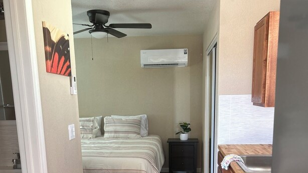 A compact room with a bed, a small bedside table with a plant, a ceiling fan, and a kitchenette with a refrigerator and wooden cabinets. - Wild West Cabin Suite (Quartzsite)