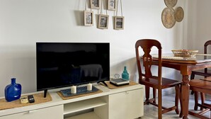A living room with a flat-screen TV, wooden dining table, chairs, and a wall-mounted picture frame with photos. - Apartamento Las Palmeras - Ole Solutions (Jerez de la Frontera)