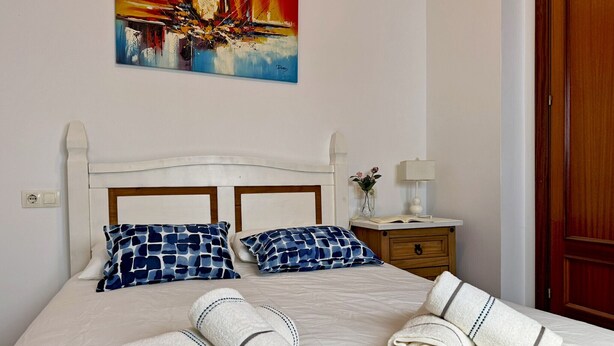 A neatly made bed with two pillows, a bedside table with a lamp, and a painting on the wall. - Apartamento Las Palmeras - Ole Solutions (Jerez de la Frontera)
