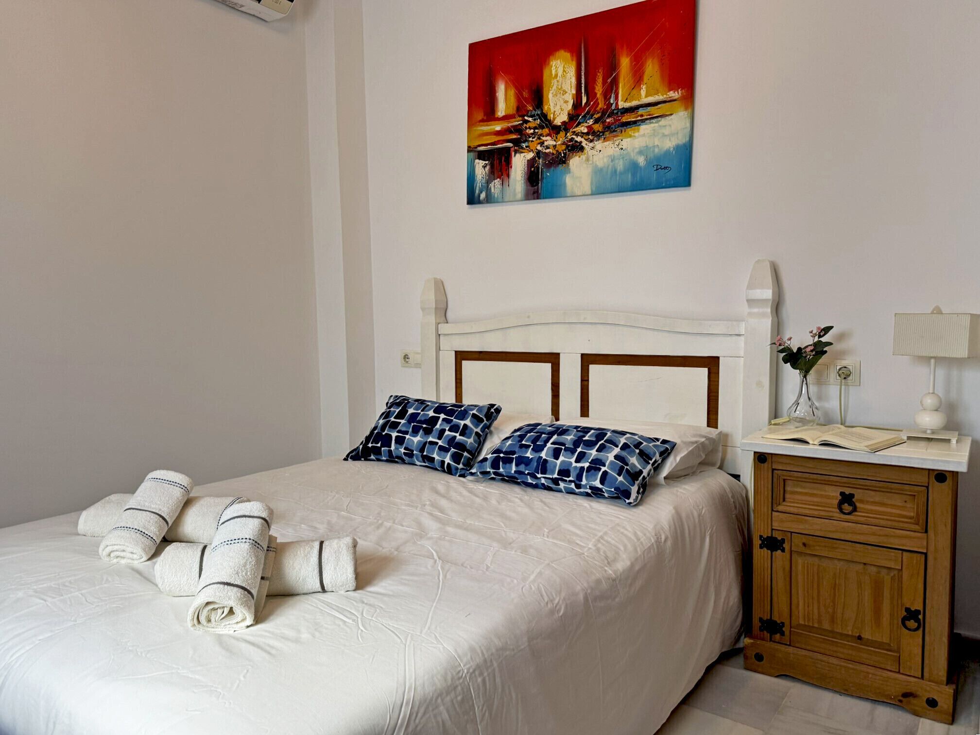 A neatly made bed with white linens and blue patterned pillows, a wooden nightstand with a lamp, and a painting on the wall.