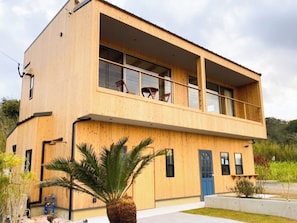 Une maison moderne à deux étages, en bois, dotée d’un balcon, d’une porte bleue et d’un petit jardin agrémenté d’un palmier.