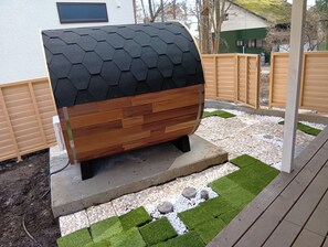Una parrilla de barbacoa moderna al aire libre con techo hexagonal, rodeada por una terraza de madera y un jardín con guijarros blancos y césped verde.