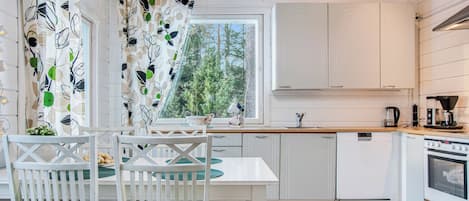 Eine moderne Küche mit weißen Schränken, einem Esstisch mit Stühlen, einem Fenster mit Blick auf Bäume und einem grünen Teppichboden.