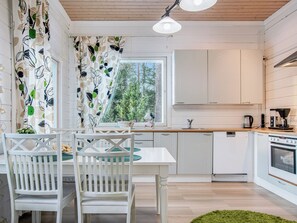 A modern kitchen with white cabinetry, a dining table with chairs, a window with a view of trees, and a green rug on the floor. - Mänty-linna by Interhome (Kihniö)