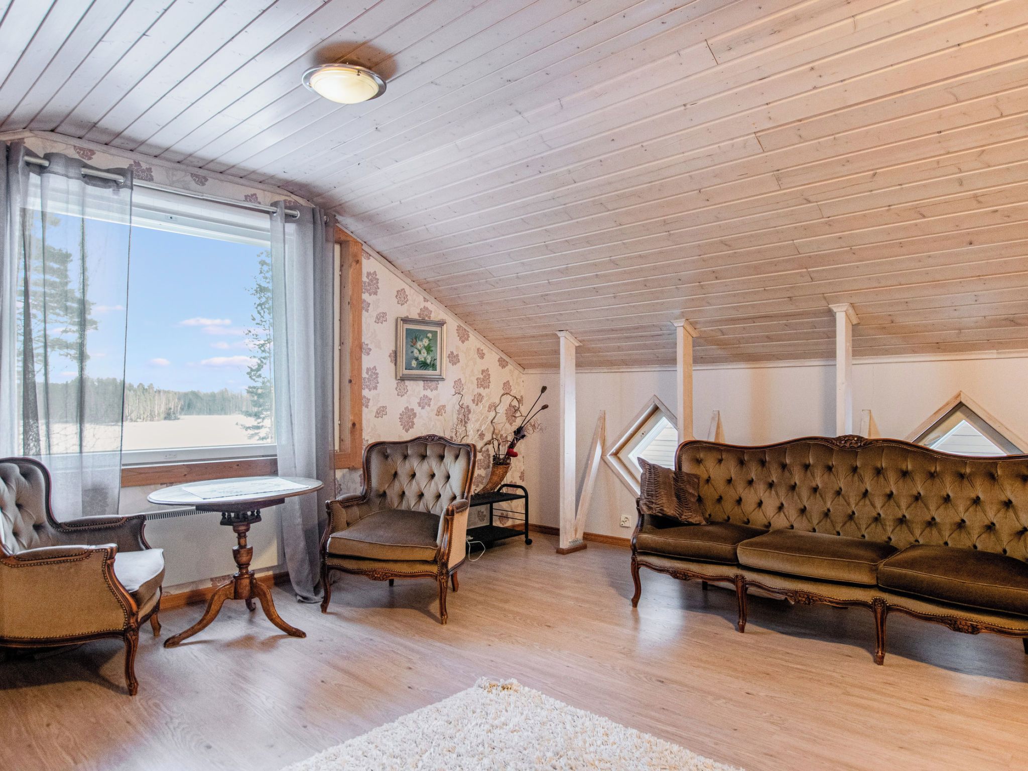 A cozy room with a wooden ceiling, a large window, a sofa, two chairs, and a small table.