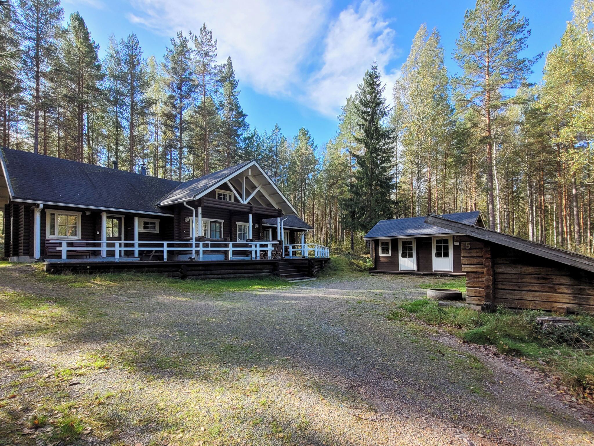 Mökki, jossa on kuisti ja erillinen rakennus, ja koko komeus sijaitsee metsän ympäröimänä.