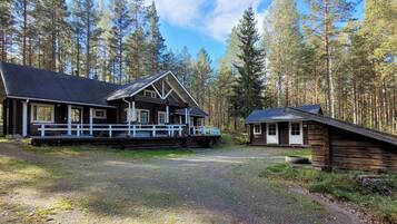 Mökki, jossa on kuisti ja erillinen rakennus, ja koko komeus sijaitsee metsän ympäröimänä.
