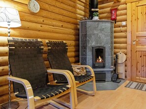 L’intérieur d’une cabane en rondins, avec une cheminée en pierre, deux fauteuils en osier, une horloge et une lampe.