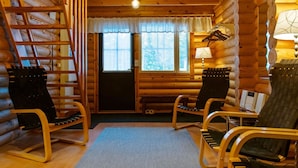 Interior de uma cabana de madeira com soalho de madeira, um tapete, duas cadeiras de balanço, uma mesa, um abajur e uma janela com cortinas.